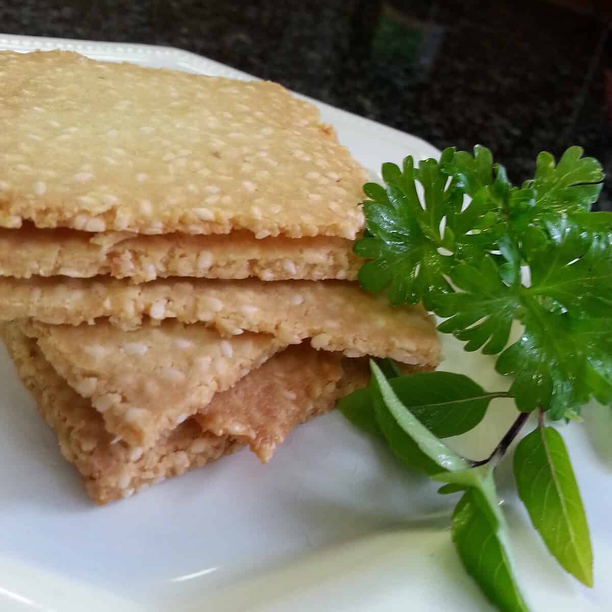 A stack of rectangular sesame crackers, perfect for keto weight loss, sits on a white plate garnished with a sprig of fresh green herbs. The blurred background draws focus to the healthy crackers and vibrant garnish.