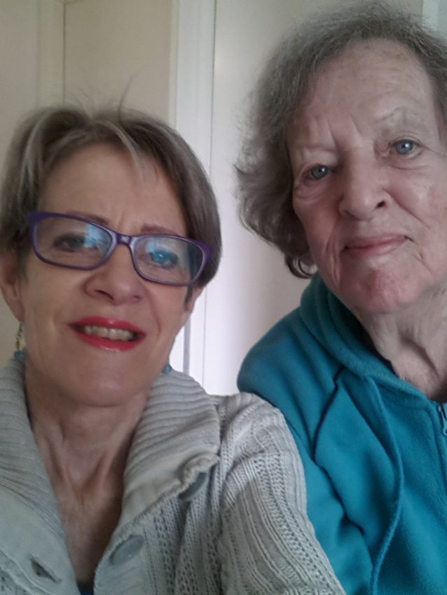 Two older women are posing for a selfie indoors, both smiling gently at the camera. One in glasses and red lipstick, the other in a blue hoodie, sharing their happiness after discovering keto weight loss success together.