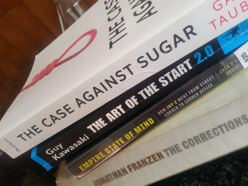 A close-up of a stack of books including The Case Against Sugar—perfect for anyone interested in keto weight loss—alongside The Art of the Start 2.0, Empire State of Mind, and The Corrections on a dark surface.