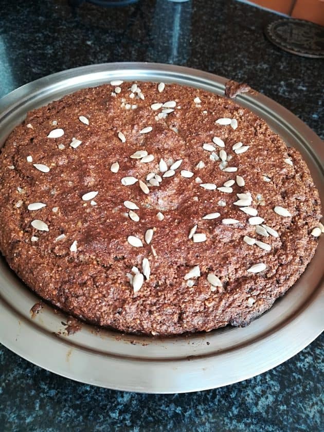 A round, homemade brown bread for keto weight loss, topped with sunflower seeds, sits on a metal plate on a dark countertop. The bread has a rustic, dense texture and a slightly cracked surface.