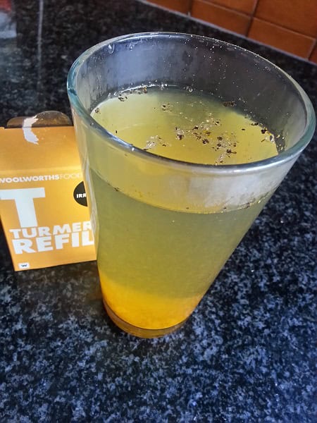 A glass of yellow turmeric drink, perfect for keto weight loss, sits on a dark countertop with spices floating on top. A box labeled Turmeric Refill is partially visible behind the glass.