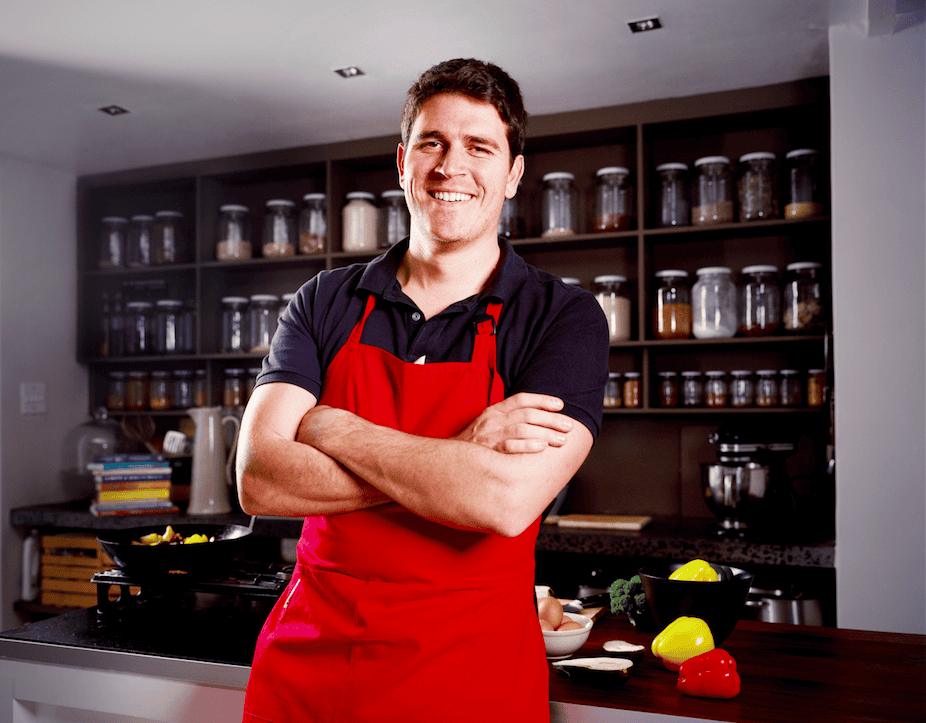 A man wearing a red apron stands in a modern kitchen with his arms crossed, smiling. Behind him are shelves filled with jars, and in front are fresh vegetables and keto weight loss ingredients on the counter.