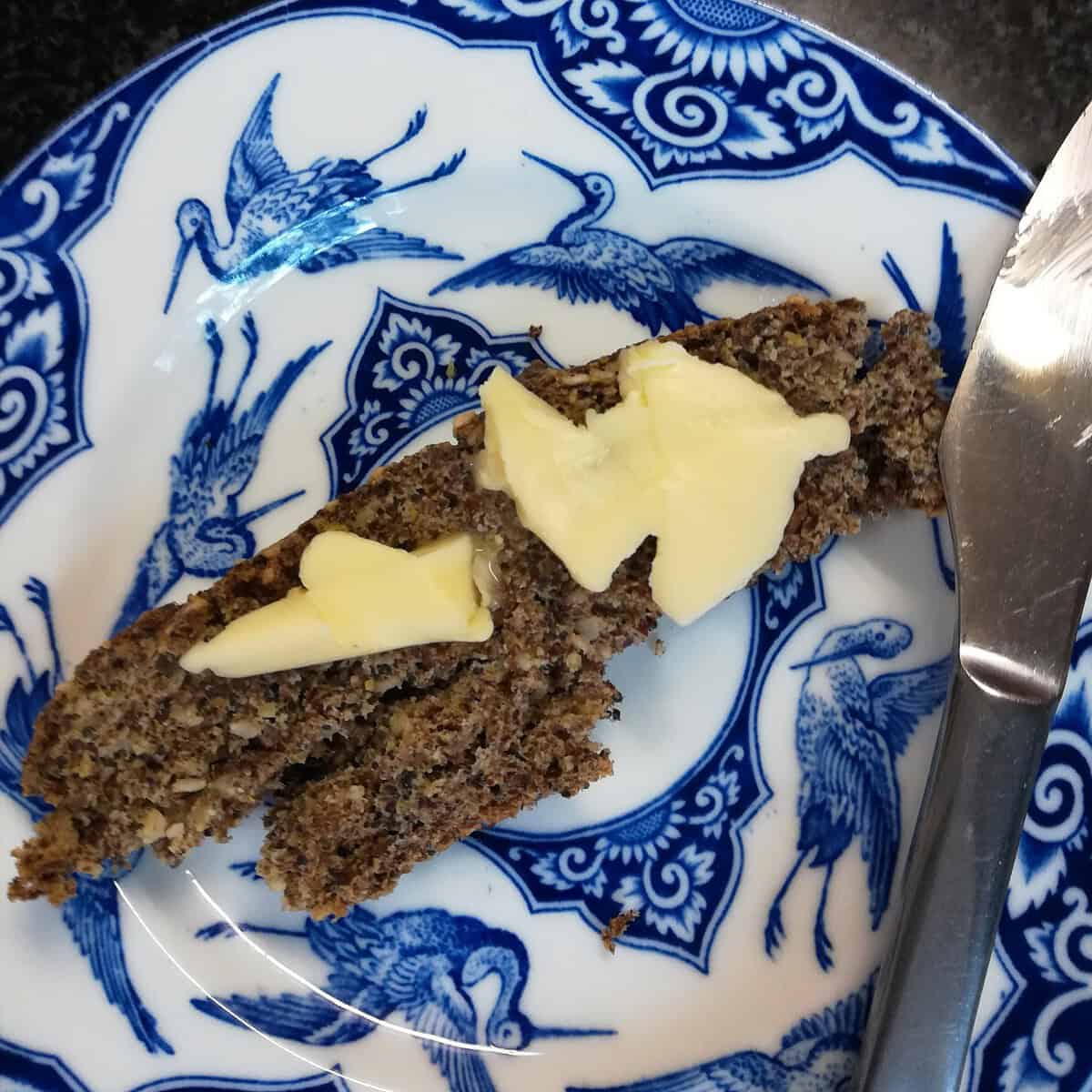 A slice of dark bread with several pieces of butter on top, served on a blue and white plate decorated with bird designs—an ideal treat for those interested in keto weight loss. A butter knife rests on the right side of the plate.