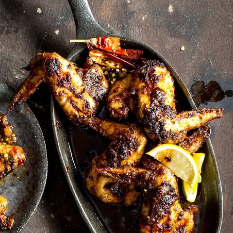 Grilled chicken wings with a dark, charred seasoning are served in a black dish, garnished with lemon wedges and dried red chilies—a delicious keto weight loss option on a rustic dark background.