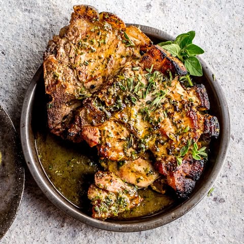 Grilled, herb-marinated pork chops served in a round metal dish, garnished with fresh green herbs and drizzled with a flavorful sauce—a keto weight loss-friendly option on a gray stone surface.