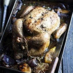 A whole roasted chicken sits in a pan surrounded by cooked vegetables and herbs, with golden crispy skin—an ideal keto weight loss meal with delicious juices pooling at the bottom.