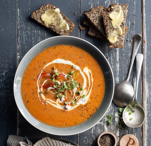 A bowl of creamy tomato soup topped with microgreens and a swirl of cream, perfect for keto weight loss, served on a dark wooden table with buttered slices of seeded bread, spoons, and a pepper shaker nearby.