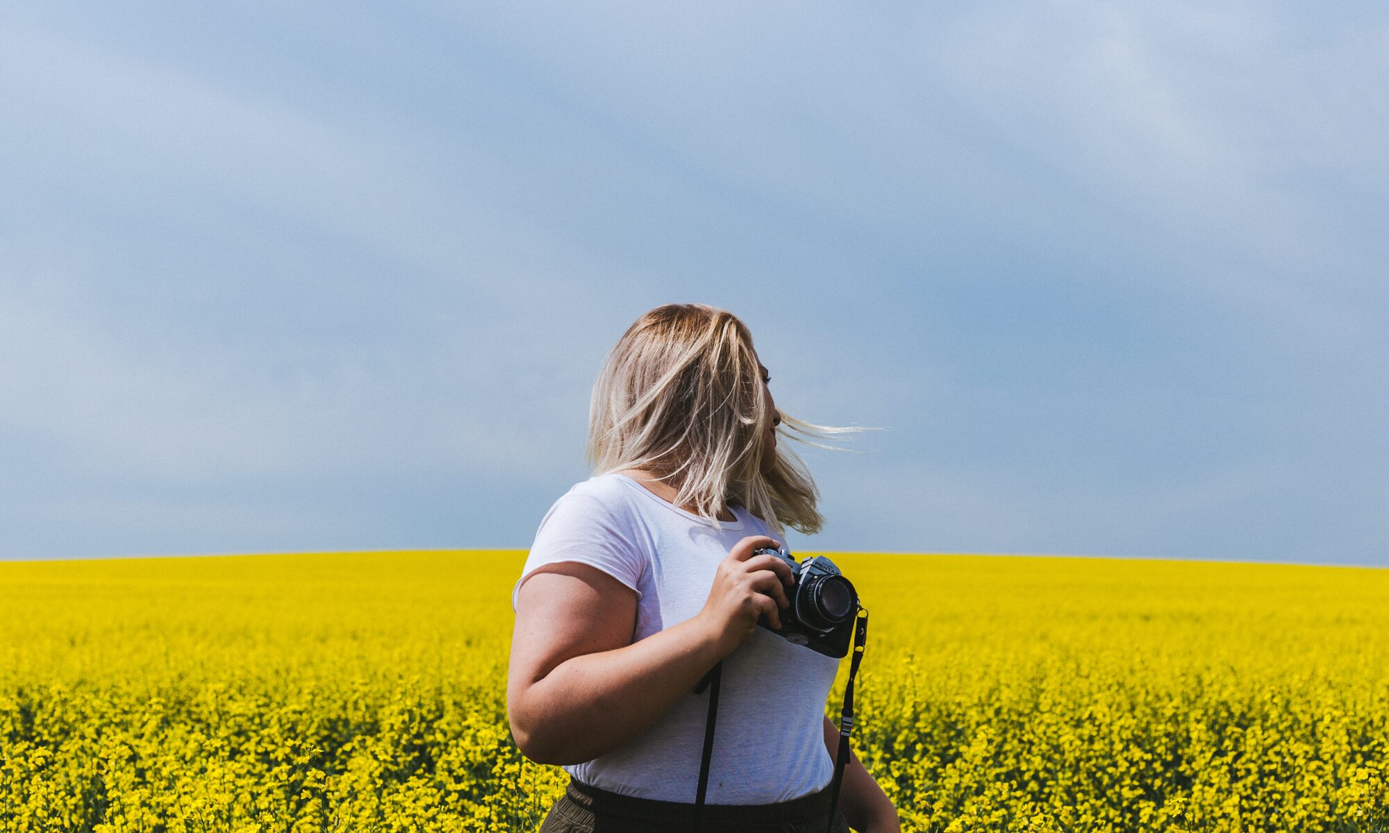 A person with blonde hair, wearing a white t-shirt, stands in a bright yellow canola field under a blue sky, holding a camera. Their confident stance suggests the positive boost that comes with successful keto weight loss.