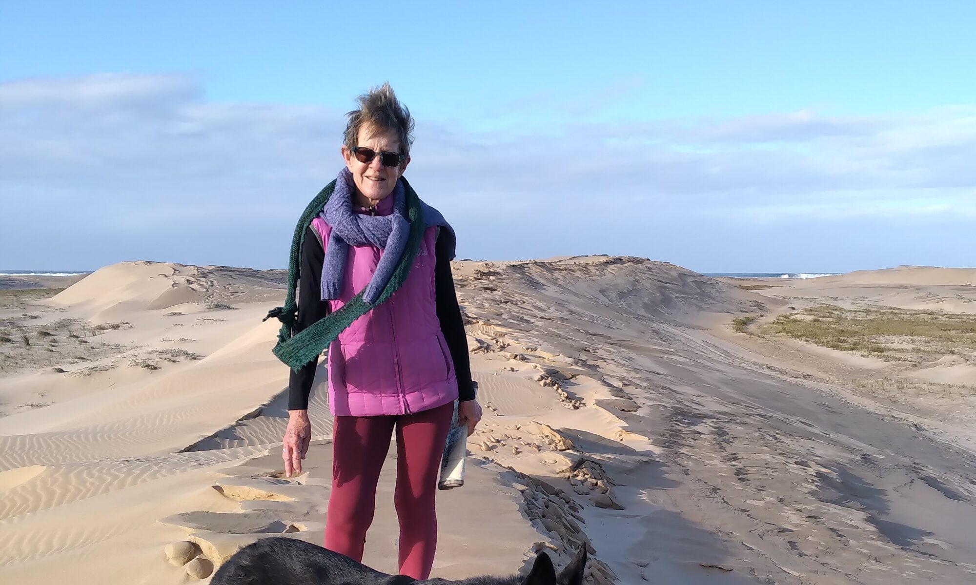 An older woman wearing sunglasses and colorful clothes stands on sandy dunes under a clear sky, smiling, with her black and white dog in the foreground—a vibrant scene that radiates energy and confidence, reflecting her keto weight loss journey.