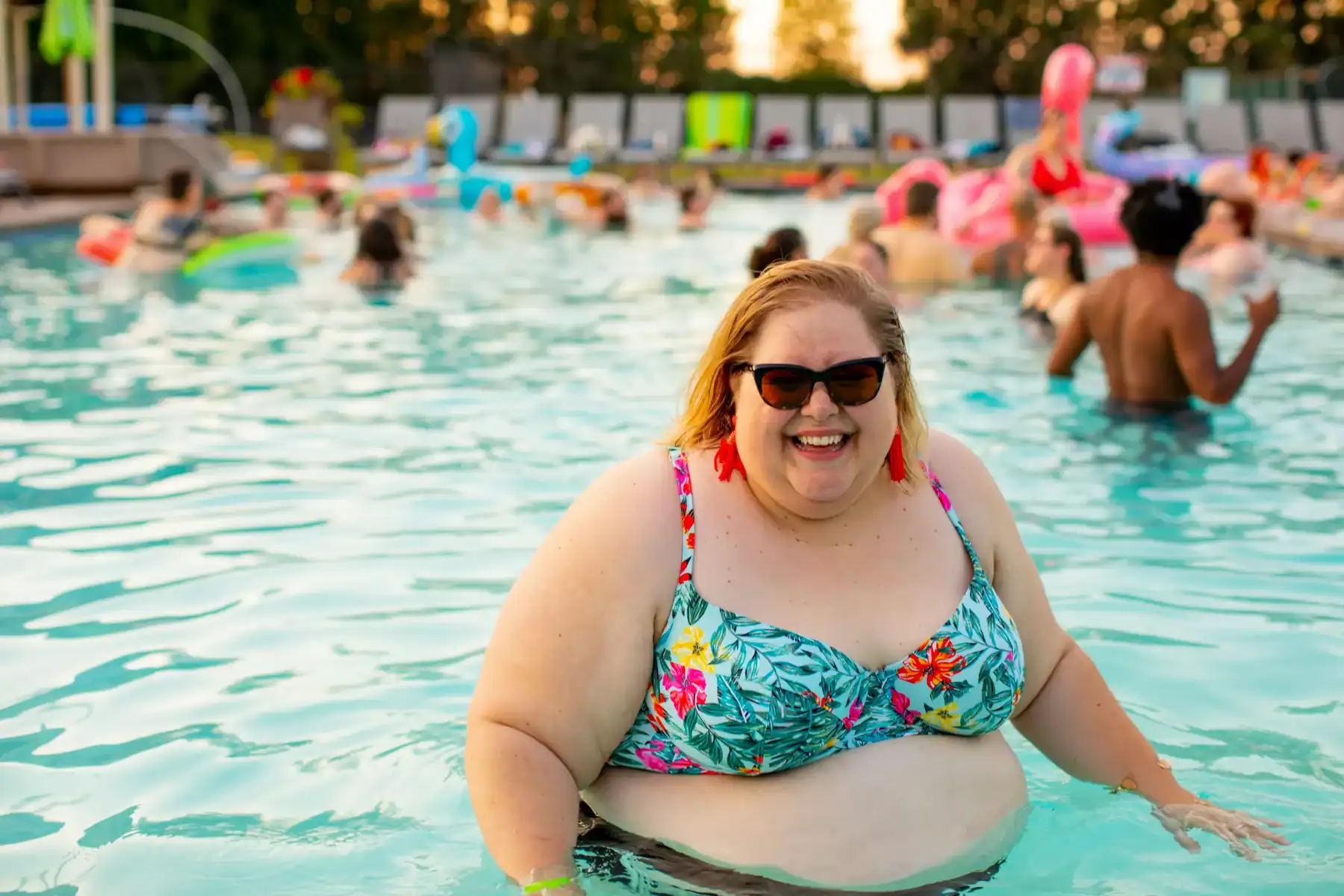 A smiling woman in a floral bikini and sunglasses stands in a busy outdoor swimming pool, radiating confidence despite recent weight gain after GLP-1, with people and pool floats in the background on a sunny day.