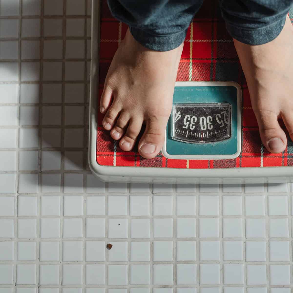 A person standing barefoot on a red and white analog bathroom scale, placed on a white tiled floor. Only the feet and lower legs are visible, subtly hinting at why the scale is misleading when it comes to assessing true health.