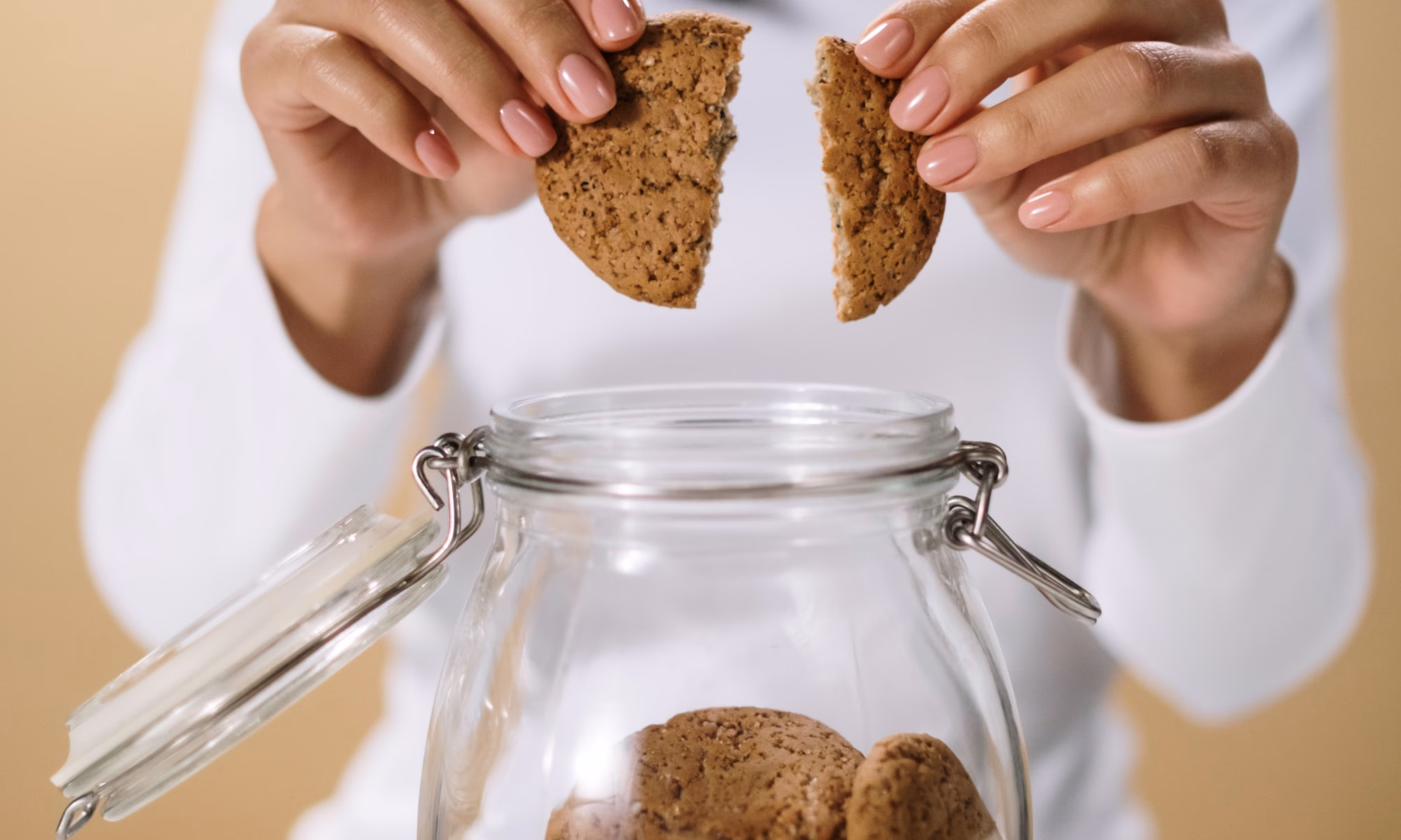 A person in a white shirt breaks a cookie in half over a glass jar filled with cookies, showing they just can’t stop eating sugar.
